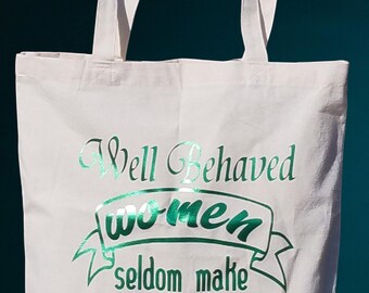 May include: Cream-coloured canvas tote bag featuring the quote "Well Behaved women seldom make HISTORY" in shimmering green text. The bag has two long handles and is suitable for carrying items.