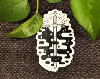 May include: Black and white sticker featuring a sword design. The sword is centered, with a detailed hilt and blade. The background has a celestial theme with stars and cloud-like shapes. Text at the bottom reads "@JULIAJODESIGNS".