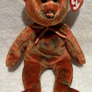 Può includere: Un orsacchiotto di peluche con un motivo tie-dye arancione e marrone, dotato di occhi in plastica nera e un naso con il logo Mastercard. Un nastro a strisce nere, rosse e gialle è legato al collo e un'etichetta Ty rossa a forma di cuore.