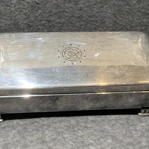 Puede incluir: Una caja rectangular de metal plateado con tapa abatible y cuatro pies pequeños escalonados. El centro de la tapa presenta un emblema circular con una estrella, un símbolo central y el texto "ADOTERAPIA/MULTITERAPIA-FANTASIA" y "1991-1992 HUNDRED CLUB".