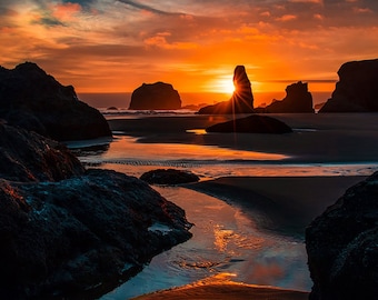 May include: A dramatic sunset over a rocky beach with the sun setting behind a tall rock formation. The sky is a vibrant orange and red, and the water is a deep blue. The rocks are silhouetted against the sky.