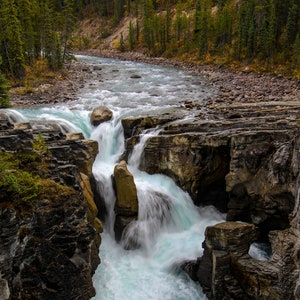 May include: A cascading waterfall flows through a rocky canyon, creating a dramatic scene of nature's power. The water is a vibrant turquoise color, and the rocks are a mix of gray and brown.