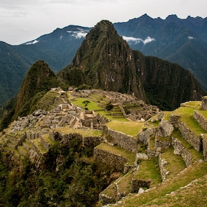 May include: An aerial view of Machu Picchu, an ancient Incan city located in the Andes Mountains of Peru. The ruins are built on a series of terraces and are surrounded by lush green vegetation.