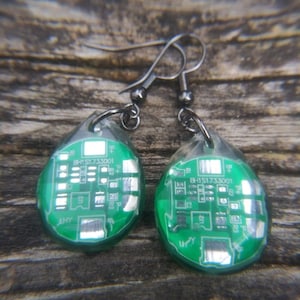 May include: A pair of green and silver earrings with a circuit board design. The earrings are made of resin and have a teardrop shape. The earrings are attached to silver hooks.