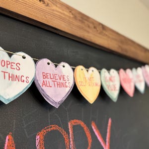May include: A string of paper hearts with different colors and phrases, including "hopes all things" and "believes all things", hanging on a black chalkboard.