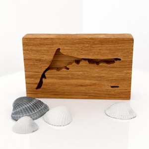 Könnte beinhalten: Ein Holzblock mit einem Ausschnitt einer Küstenlinie. Das Holz hat eine natürliche Maserung. Mehrere Muscheln in Grau- und Weißtönen sind vor dem Block angeordnet, was ein Küstenmotiv ergibt.