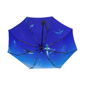 May include: Open umbrella with a deep blue background featuring a school of sharks swimming in the ocean. The umbrella has a black handle and frame, and the sharks are white and gray.