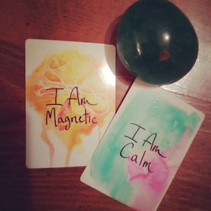 以下が含まれることがあります： 水彩画の背景を持つ肯定的なカードが2枚。最初のカードは黄色とオレンジで、「私は磁力的です」というテキストが書かれています。2枚目のカードは緑とピンクで、「私は落ち着いています」というテキストが書かれています。