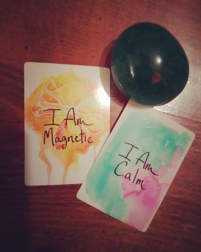 以下が含まれることがあります： 水彩画の背景を持つ肯定的なカードが2枚。最初のカードは黄色とオレンジで、「私は磁力的です」というテキストが書かれています。2枚目のカードは緑とピンクで、「私は落ち着いています」というテキストが書かれています。