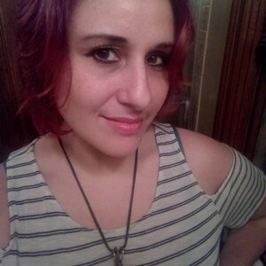 以下が含まれることがあります： ピンクの髪の女性は、ストライプのオフショルダーシャツを着て、長い黒いコードネックレスに大きな暗いペンダントをつけています。