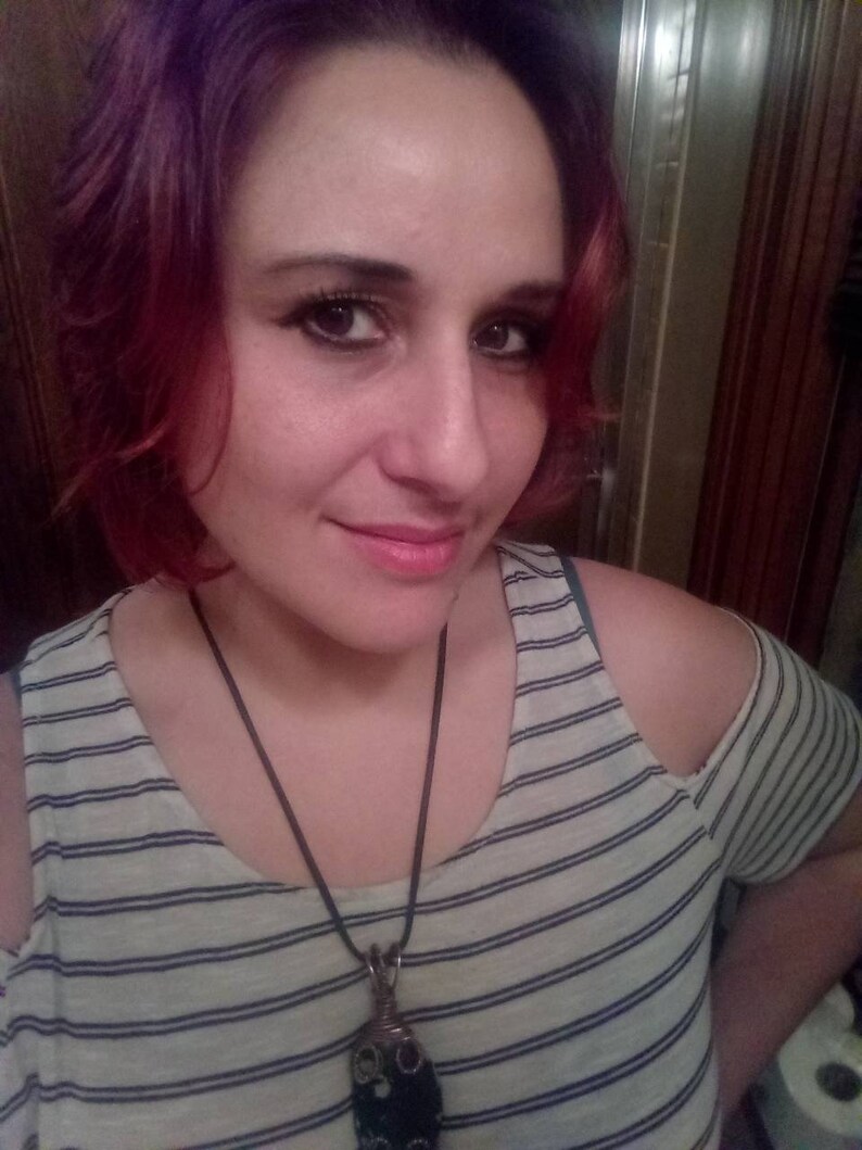 以下が含まれることがあります： ピンクの髪の女性は、ストライプのオフショルダーシャツを着て、長い黒いコードネックレスに大きな暗いペンダントをつけています。