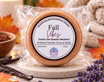May include: A round, tan-colored aromatherapy bath bomb and shower steamer in clear plastic packaging. The label reads Fall Vibes, Smells Like Sweater Weather!, All Natural Lavender, Cocoa, & Vanilla, Made in USA, and 4.5 OZ with a lotus logo.
