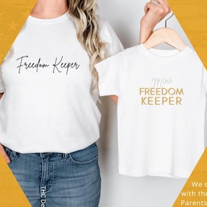 May include: White t-shirts with the text "Freedom Keeper" and "Mini Freedom Keeper" in a script font. The shirts are displayed on a yellow background with text that reads "We don't coparent with the government."