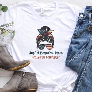 May include: White t-shirt featuring a graphic of a woman with sunglasses and a patriotic headband. The text reads "Just A Regular Mom Raising Patriots" in a mix of blue and red colors. The shirt is paired with blue jeans and gray boots.
