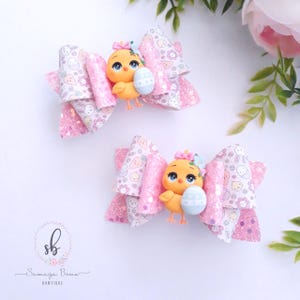 May include: Two Easter-themed hair bows. Each bow features a yellow chick holding a blue and white egg. The bows have layers of pink and white patterned fabric with glitter accents. The patterns include Easter eggs, carrots, and bunnies.