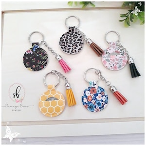 May include: A collection of keychains with decorative round tags. The tags feature various patterns, including floral, leopard print, and honeycomb designs. Each keychain has a tassel and a silver-colored chain and ring. The brand name "Samaga Bowtique" is visible.