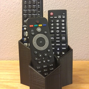 May include: A black 3D printed remote control organizer with three slots for remotes. The organizer is holding three different black remotes with buttons.