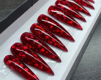 Op de afbeelding: Een set lange, stiletto-vormige valse nagels. De nagels zijn dieprood met ingebedde glitter, gerangschikt in een doos. De glitter vangt het licht op en creëert een sprankelend effect. De nagels zijn netjes gerangschikt in een witte doos met een zwarte rand.
