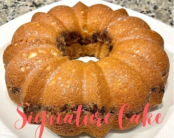 Könnte beinhalten: Ein goldbrauner Bundtkuchen mit einer leichten Puderzuckerbestäubung. Der Kuchen befindet sich auf einem weißen Teller und der Text "Signature Cake" ist in roter Kursivschrift unter dem Kuchen geschrieben.