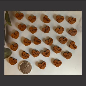 May include: A scattering of small, wooden heart-shaped embellishments, each engraved with the word "Love". The hearts are a warm brown colour and scattered across a white surface. A coin is also present.