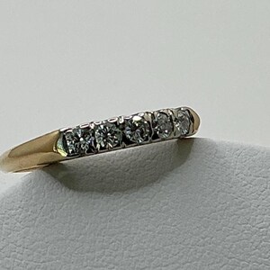 May include: A gold band ring with five clear, round-cut gemstones set in a row. The ring has a classic design, with a polished gold finish and a simple, elegant style. The gemstones sparkle brightly.