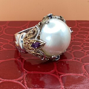 May include: A silver and gold ring with a large white pearl and purple gemstones. The ring is set on a red background.