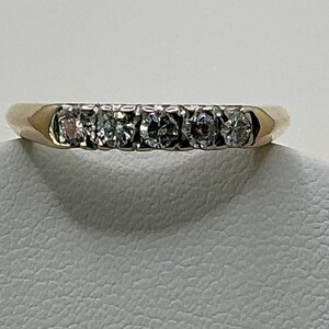 May include: A gold-toned ring with five small, clear gemstones set in a row. The ring band is smooth and polished, with a slight taper towards the gemstones. The gemstones are round and evenly spaced.