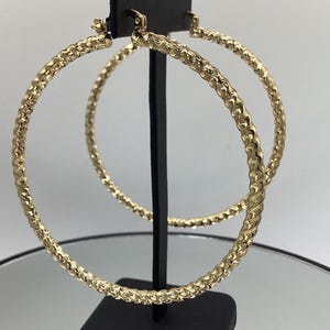 Puede incluir: Un par de pendientes de aro grandes, de color dorado, con un diseño texturizado y ondulado. Los pendientes se exhiben en un soporte negro sobre un fondo neutro. Los aros son redondos y tienen un acabado brillante.