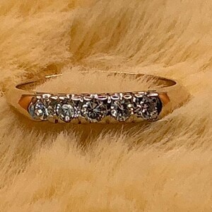 May include: A gold band ring featuring five round-cut diamonds. The diamonds are set in a row across the top of the ring. The ring is set against a soft, textured background.