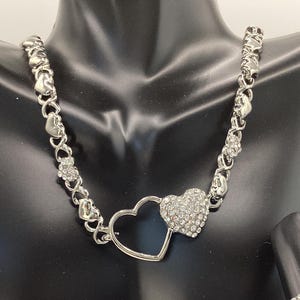 May include: A silver necklace with a double heart pendant. The hearts are connected and have a pave setting of clear crystals. The chain is made of small linked hearts.