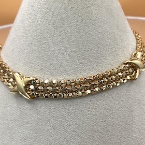 Puede incluir: Un brazalete de cadena de oro con un diseño tejido y un cierre en forma de X.