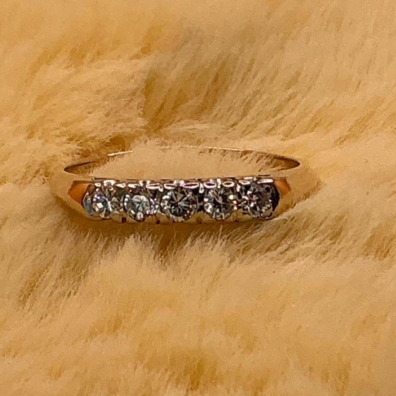 May include: A gold band ring featuring five round-cut diamonds. The diamonds are set in a row across the top of the ring. The ring is set against a soft, textured background.