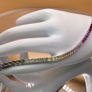 May include: A tennis bracelet with a gradient of colorful gemstones, ranging from blue to red. The bracelet is held by a white hand-shaped display, showcasing the vibrant array of colors and the jewelry's design.
