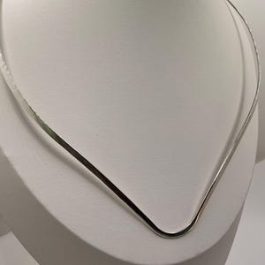 Puede incluir: Un collar plateado con diseño en forma de V, exhibido sobre un busto de maniquí blanco. El metal es liso y reflectante, captando la luz. Diseño sencillo y elegante.