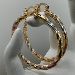 May include: A pair of large hoop earrings featuring a tri-color metal design with alternating sections of yellow, rose, and white gold tones. The surface of the hoops has a textured, ribbed pattern throughout, and each earring includes a hinged clasp.