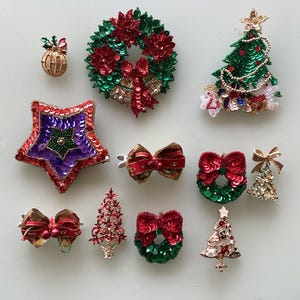 Peut inclure: Une collection de broches sur le thème de Noël avec une variété de motifs, notamment des couronnes, des étoiles, des arbres et des nœuds. Les broches sont faites de paillettes et de perles dans des tons de rouge, vert, or et violet.