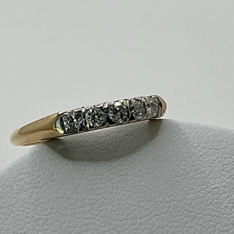 May include: A gold band ring with five clear, round-cut gemstones set in a row. The ring has a classic design, with a polished gold finish and a simple, elegant style. The gemstones sparkle brightly.