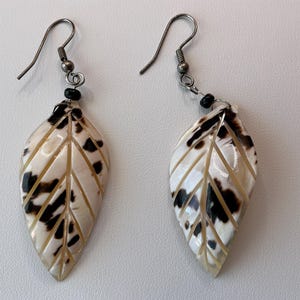 May include: A pair of leaf-shaped earrings. Each earring features a cream-colored, carved leaf design with dark brown spots and veins. The earrings have silver-colored hooks and small black beads.