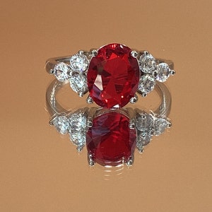 Pode incluir: Um anel de prata com uma grande gema oval vermelha cercada por gemas menores transparentes.