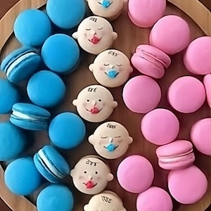 May include: A collection of blue and pink macarons decorated with baby faces. Some macarons have a blue pacifier, while others have a red mouth. The macarons are arranged in a circle on a wooden surface.