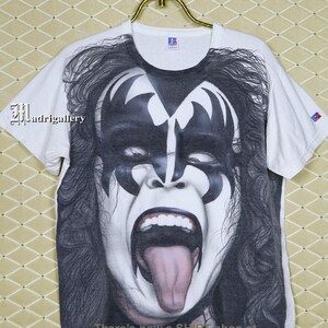 May include: White t-shirt featuring a black and white image of a person with their tongue sticking out. The image is of a rock star with makeup and a black and white design on their face. The text "There's now a Shirt shop at www.CTMadrigal.com" is printed on the bottom of the shirt.