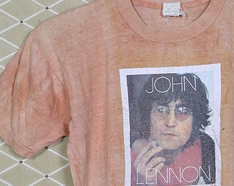 Puede incluir: Una camiseta vintage de color melocotón con una imagen descolorida de John Lennon con gafas y una camisa roja. El texto "John Lennon" está impreso debajo de la imagen.