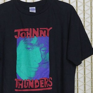 Puede incluir: Camiseta negra con un gráfico rojo y azul de Johnny Thunders, un músico. El gráfico presenta un primer plano de su rostro con el texto "JOHNNY THUNDERS" encima.