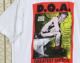 May include: White t-shirt with a red and black graphic print of a band called "D.O.A." The graphic features a man sitting on a toilet with the text "Greatest Shits" below.