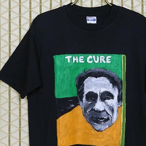 May include: Black t-shirt with a graphic print of Robert Smith, the lead singer of The Cure, in a yellow shirt and green background. The text "THE CURE" is printed in white above the image.