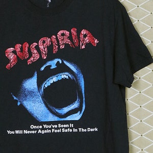 May include: Black t-shirt with a graphic print of a screaming face and the text "SUSPIRIA" in red and white. The text below the graphic reads "Once You've Seen It You Will Never Again Feel Safe In The Dark."