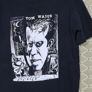 May include: Black t-shirt with a white graphic print of Tom Waits. The graphic features a black and white illustration of Tom Waits with a microphone and musical instruments.
