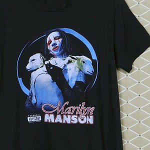 May include: Black t-shirt with a graphic of Marilyn Manson holding two white lambs. The graphic is in a circle with a blue background. The text "Marilyn Manson" is printed below the graphic. The Parental Advisory sticker is visible in the bottom left corner.