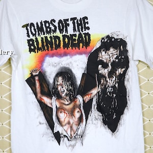 May include: White t-shirt featuring a graphic print of a woman with a skull behind her. The text "Tombs of the Blind Dead" is printed on the shirt.