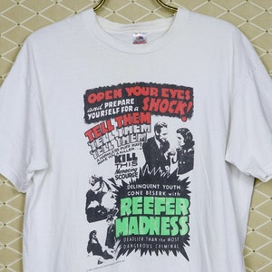 Op de afbeelding: Wit t-shirt met een grafische afbeelding die de film "Reefer Madness" promoot. Het ontwerp bevat tekst in rood en groen, samen met zwart-wit illustraties. Het t-shirt hangt aan een goudkleurige hanger.